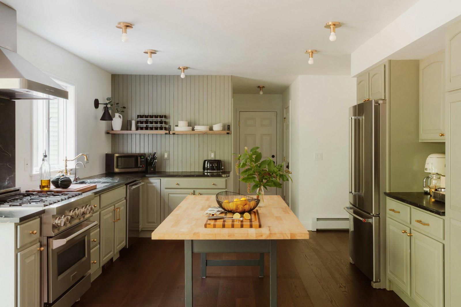 Modern sage green kitchen with a butcher block island, stainless steel appliances, and minimalist gold ceiling lights.